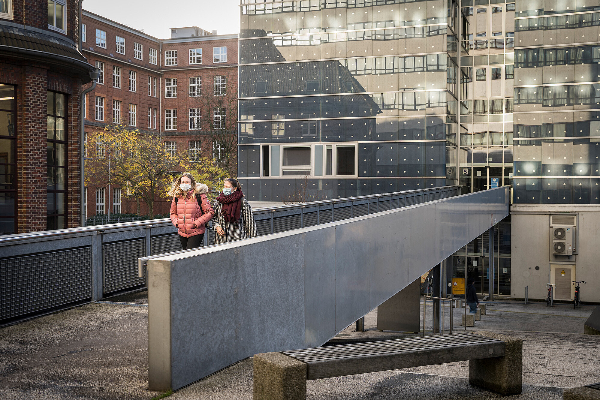 
                Zwei Studentinnen mit Maske und Winterkleidung gehen über eine Fußgängerbrücke
            