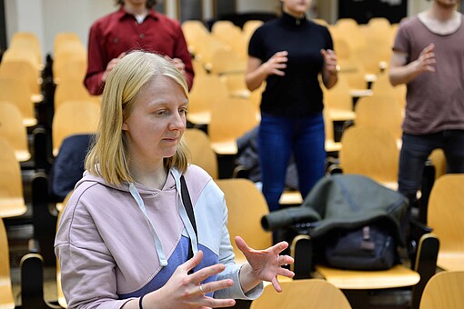 
                Studentin mit drei Studierenden im Hintergrund bei einem Workshop der Kompetenz-Werkstatt in einem Hörsaal.
            