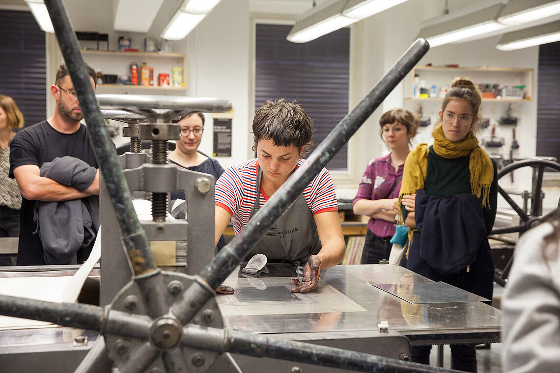 Woman standing in a workshop with others watching as she works on a litography demonstration